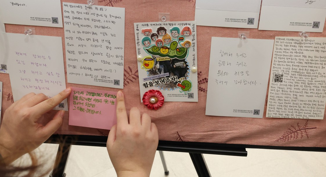 여러 색 메모지가 빼곡히 붙은 게시판에 한 사람의 손이 새 메모지를 붙이는 모습