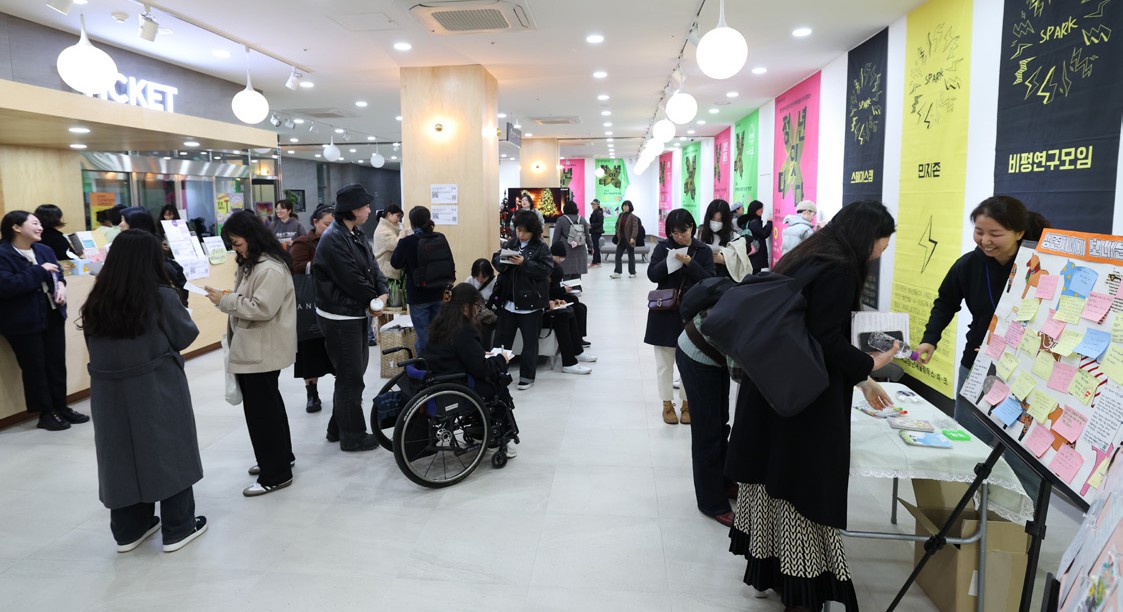 휠체어 이용자를 포함한 많은 사람들이 로비에 모여 있고, 일부는 메시지 게시판 앞에서 글을 쓰고 있는 모습